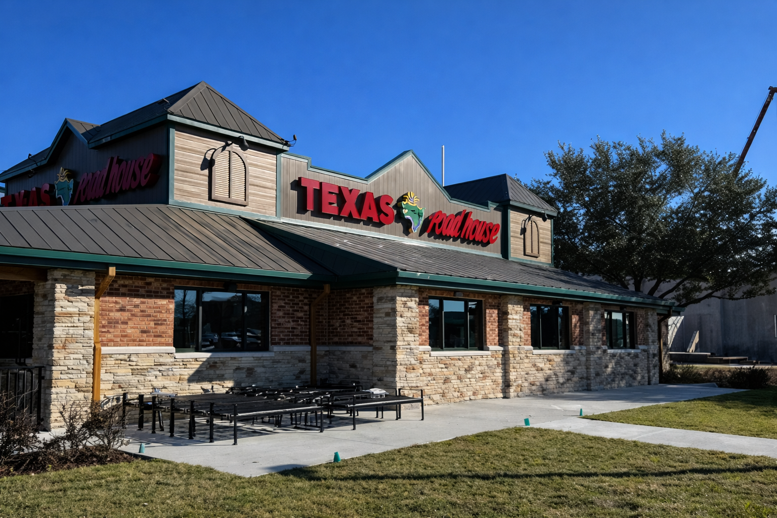 Front view of a newly constructed Texas Roadhouse in Kissimmee, FL, featuring a mix of brick and stone veneer, green metal roofing, bold red signage, and a concrete patio area ready for outdoor seating, all under a clear blue sky.