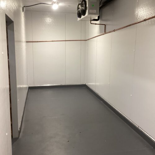 Interior view of newly constructed commercial cooler with sealed gray flooring, insulated wall panels, ceiling-mounted refrigeration unit, and finished plumbing lines at a restaurant project in Lakewood, Florida.