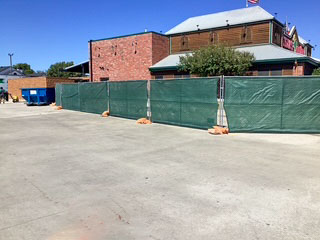 Jobsite setup for new restaurant addition in Bedford, TX—temporary green construction fencing installed in front of existing brick building to prepare for 690 sq ft dining room expansion.